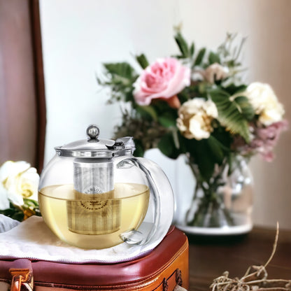 "Elegant glass teapot with a stainless steel infuser, filled with tea, placed on a leather surface. A bouquet of pink and white flowers in a vase is in the blurred background, adding a touch of sophistication and warmth to the scene."