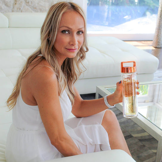 "Blonde woman with long hair wearing white attire, seated comfortably on a couch, holding a Wayfarer tea bottle and smiling at the camera."