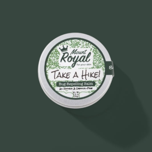 A tin of Mount Royal’s Take-a-Hike Balm on a rustic wooden surface, surrounded by natural ingredients like beeswax, calendula, and eucalyptus leaves.